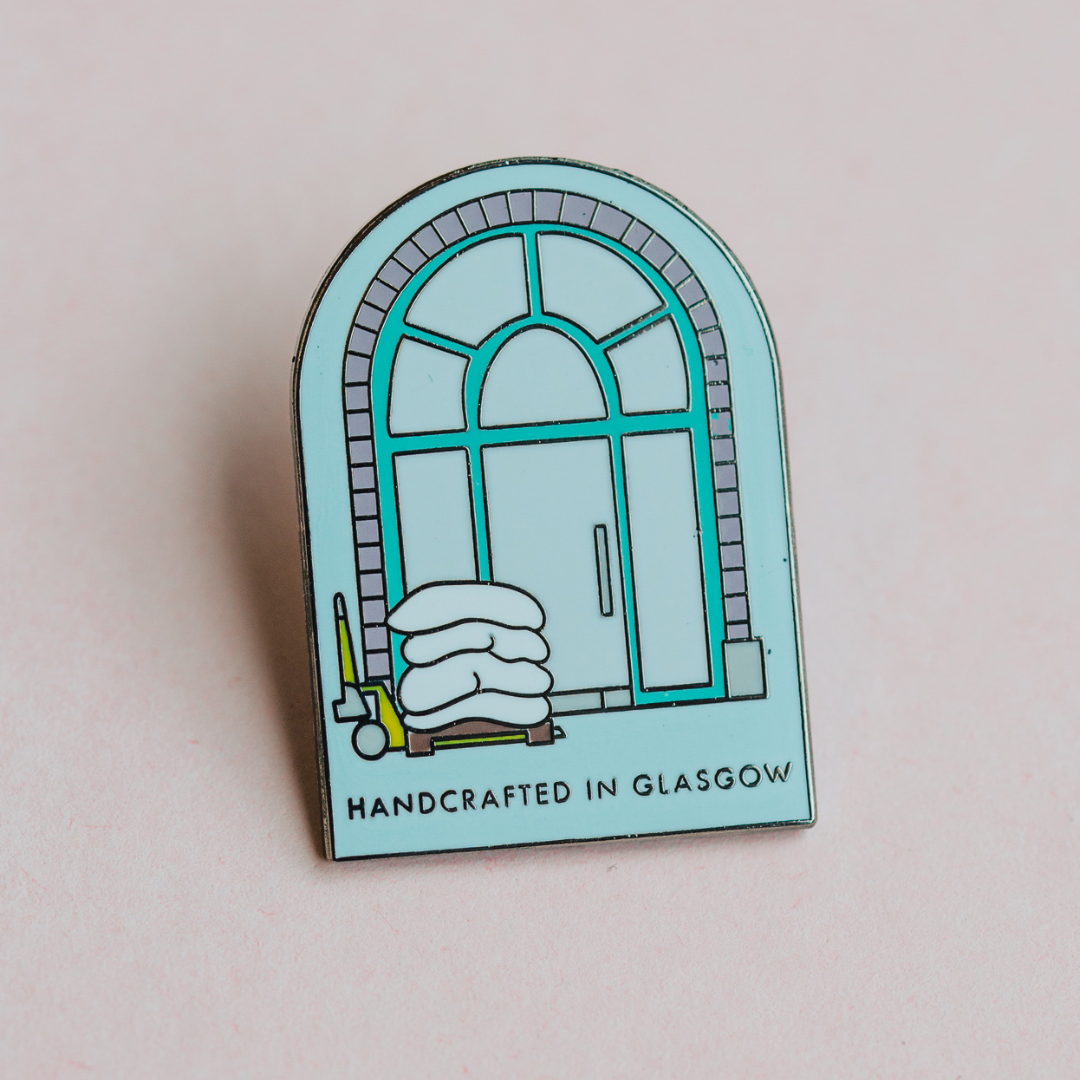 A small pin badge used by Bare Bones. It shows an ornate window in a pale blue colour. 